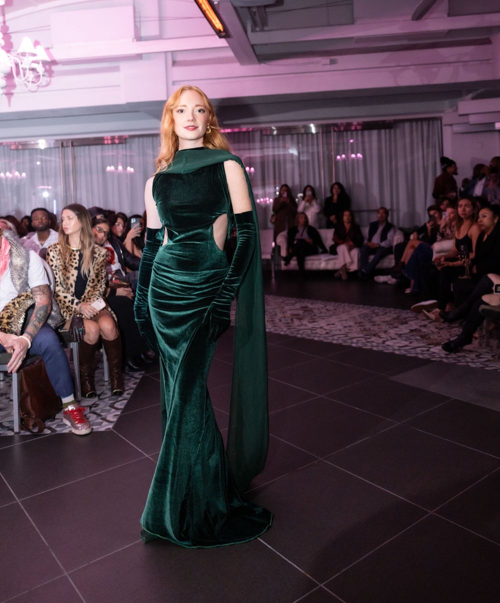 Elegant emerald velvet evening gown with matching gloves showcased on pink-lit catwalk.