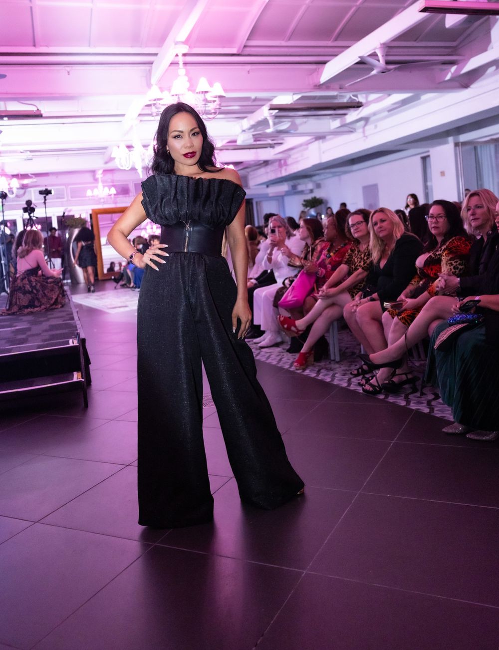 Dramatic black wide-leg jumpsuit with structured shoulders on pink-lit fashion runway.