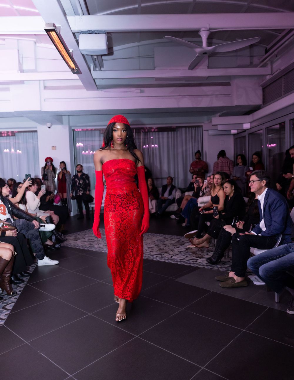 Model walks runway in stunning red lace dress and matching headpiece at indoor fashion show.