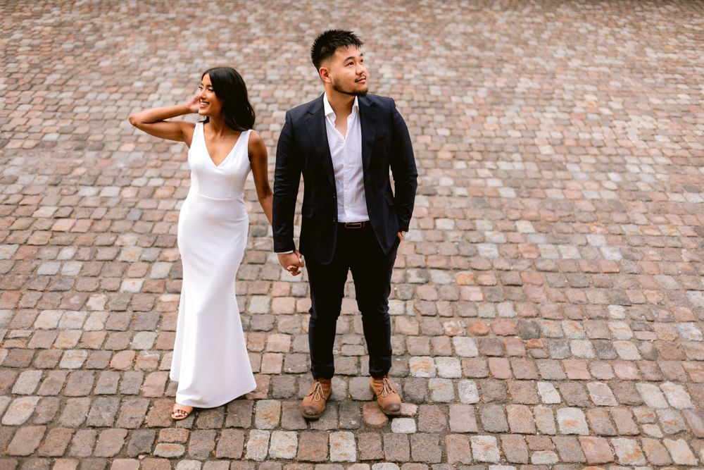 Stylish couple in formal attire posing on cobblestone street with artistic composition.