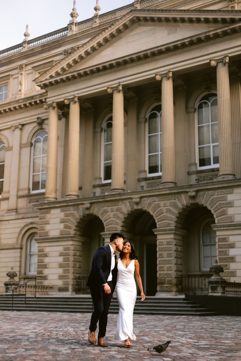 A couple shares an intimate moment in front of a grand neoclassical building with elegant columns and arched windows.