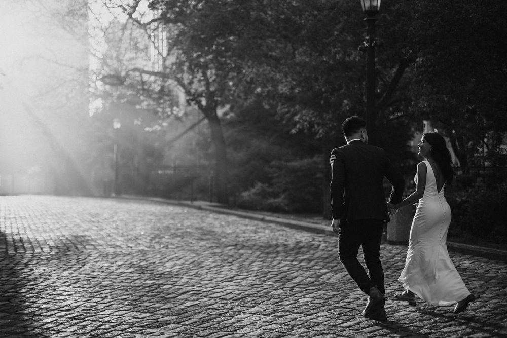 Black and white photo of couple walking on cobblestone path with sunlight streaming through trees.