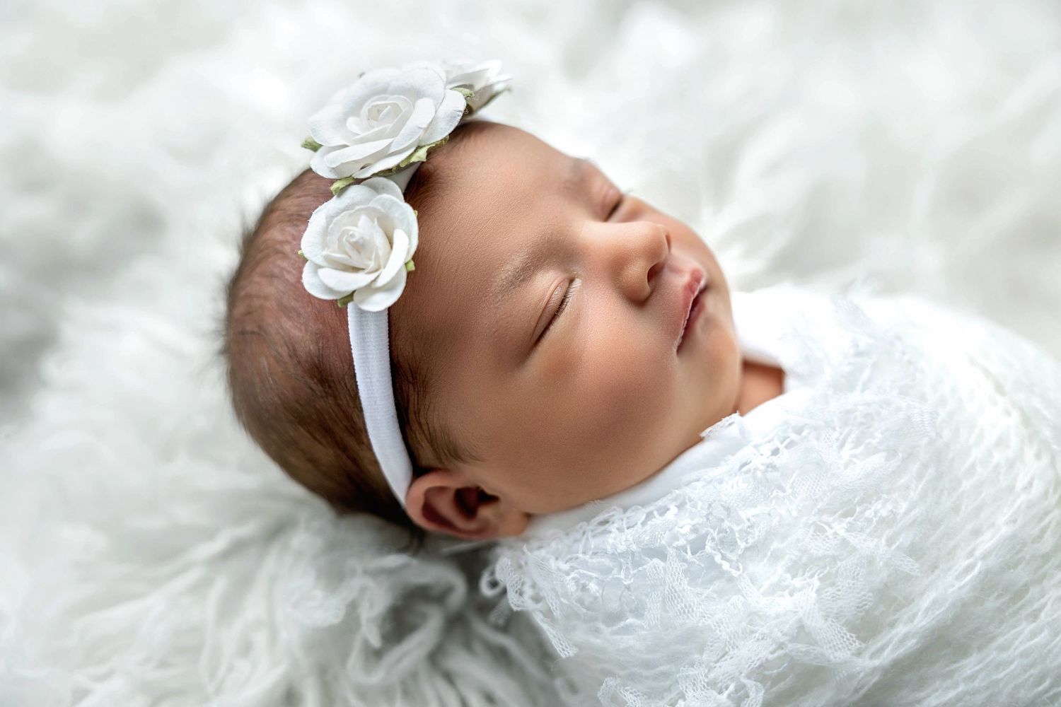 Sleeping newborn baby wearing white lace dress and floral headband rests peacefully on soft white fabric.