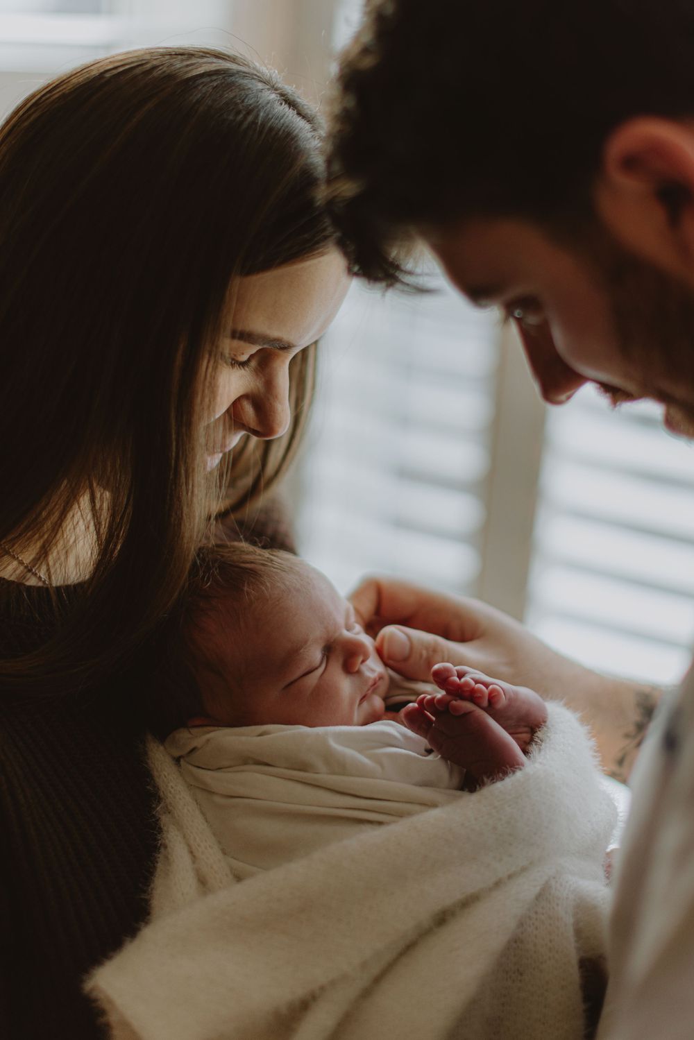 Tender moment between adults looking down at newborn wrapped in white blanket by window.