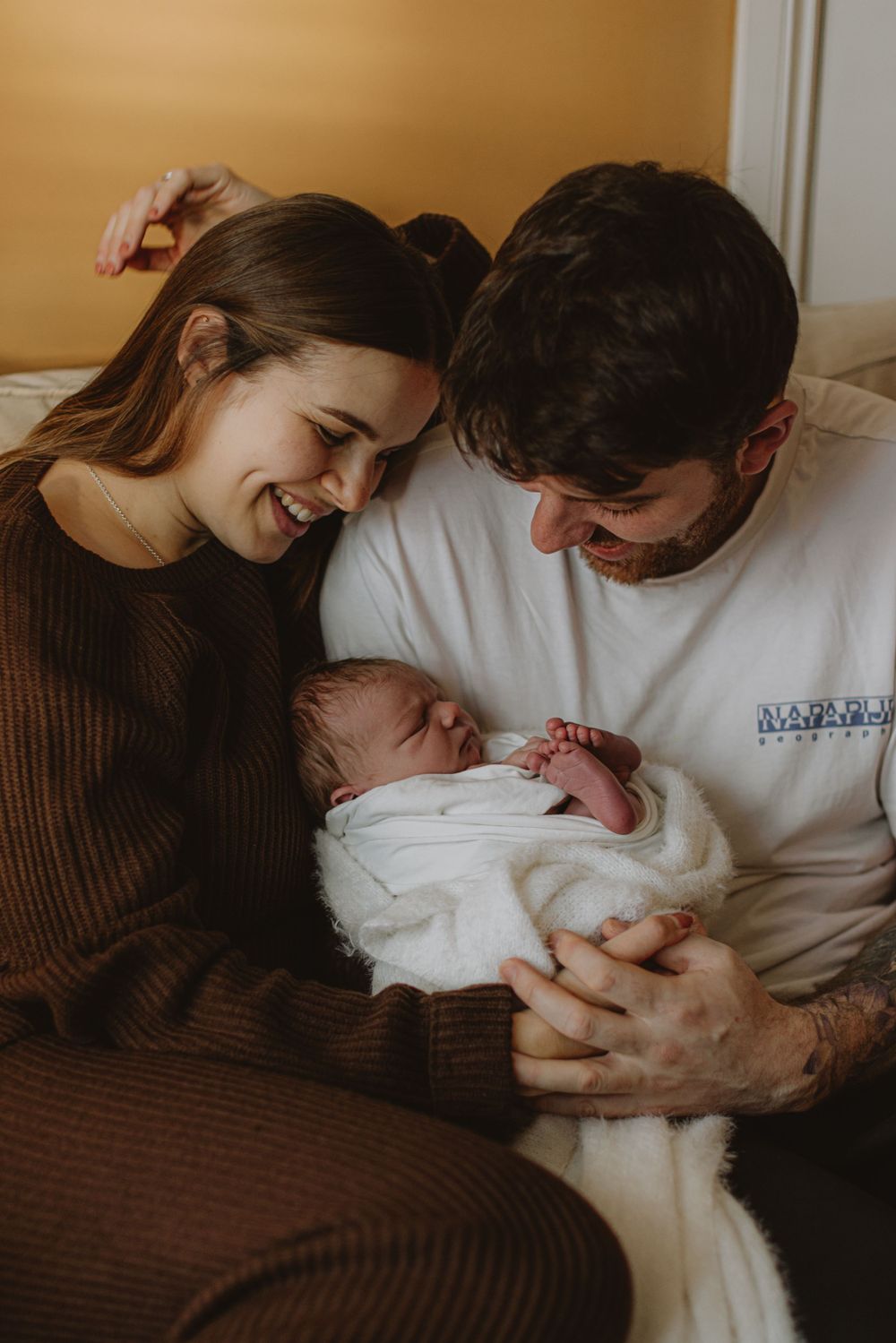 Family sharing intimate moment on brown couch while holding newborn in white blanket.