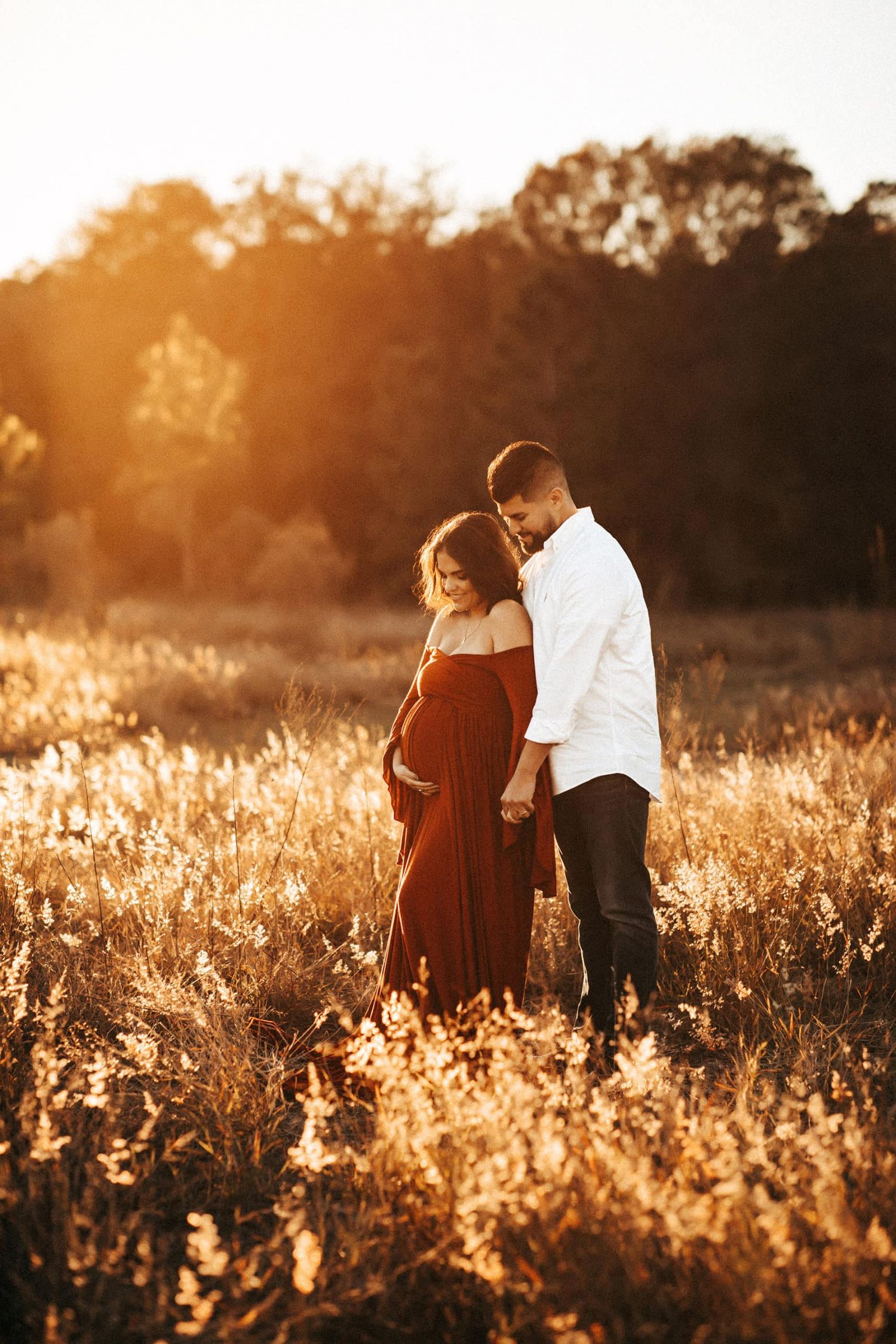 A couple embraces in a golden sunlit field during sunset, creating a warm and romantic maternity photo.