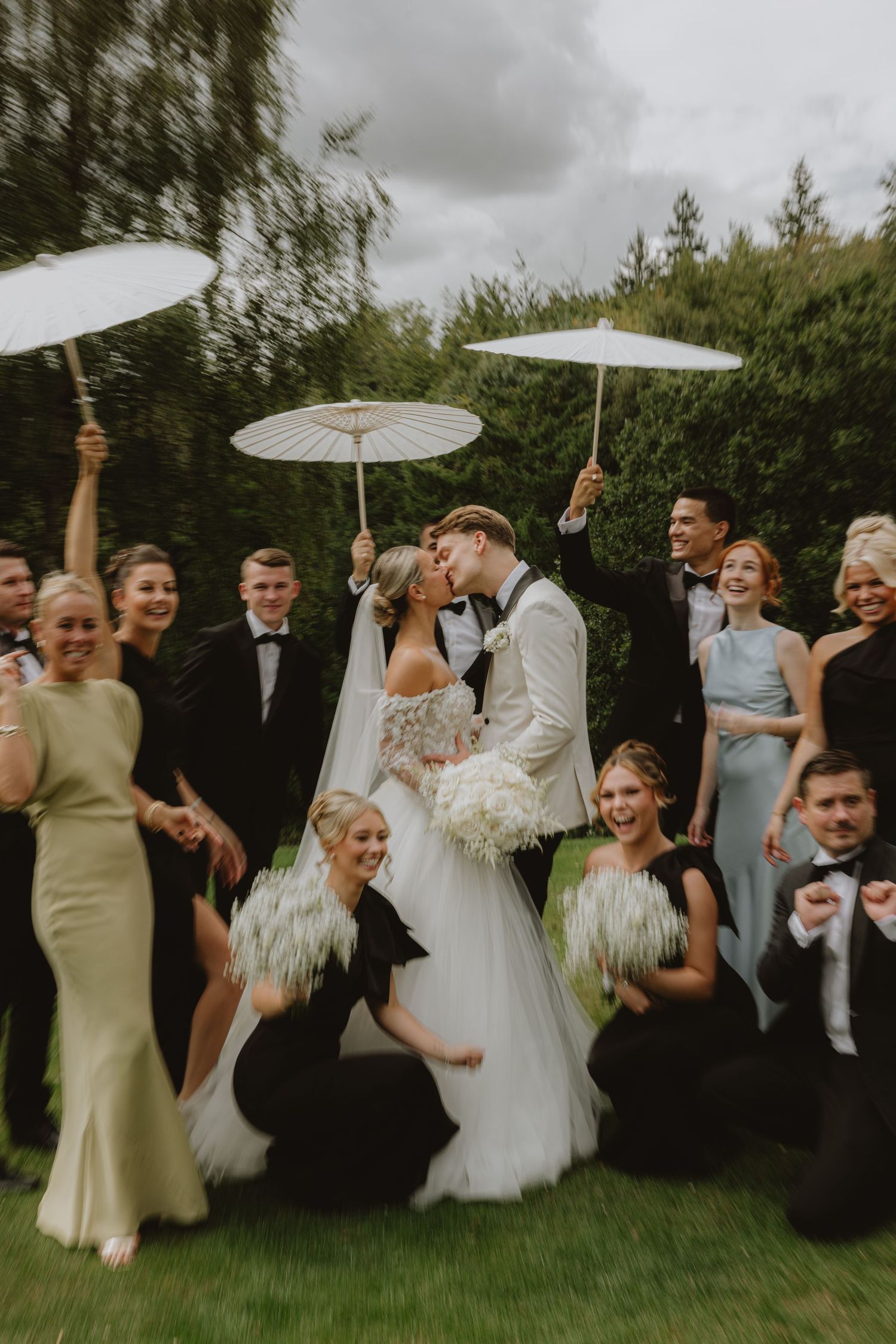 Wedding party celebrates outdoors with white parasols against a garden backdrop.