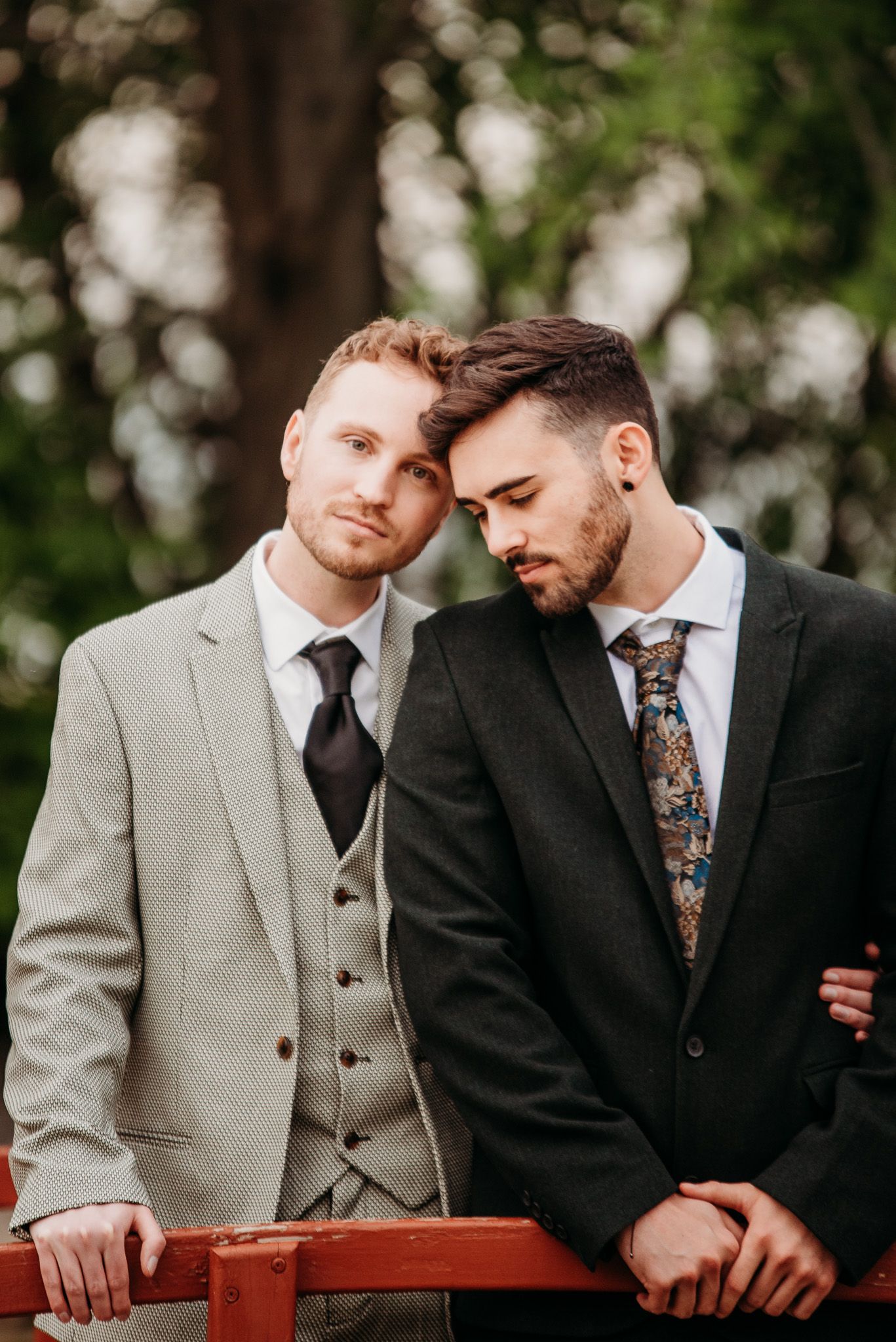 Two guests in formal suits share a tender moment against a blurred natural backdrop.