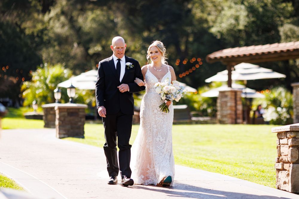 A joyful wedding couple walks along a scenic outdoor path surrounded by lush greenery and gardens.