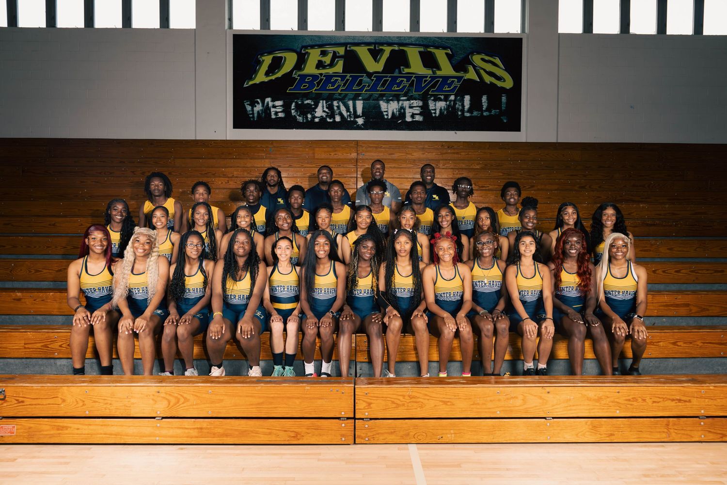 A large girls track and field team poses together in uniforms on gymnasium bleachers.