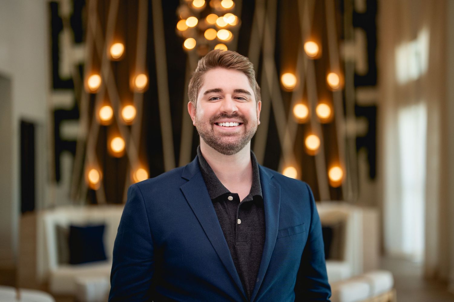 Professional portrait with person in navy blazer and burgundy shirt smiling in front of warm bokeh lighting.