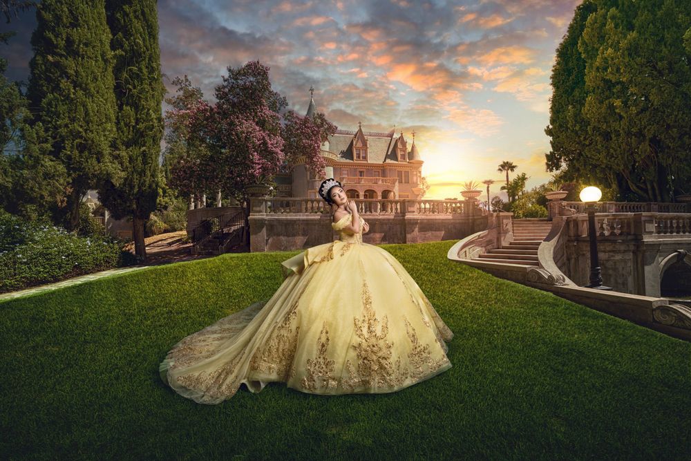 Woman in golden ball gown poses at sunset near a fairy-tale castle surrounded by lush gardens.