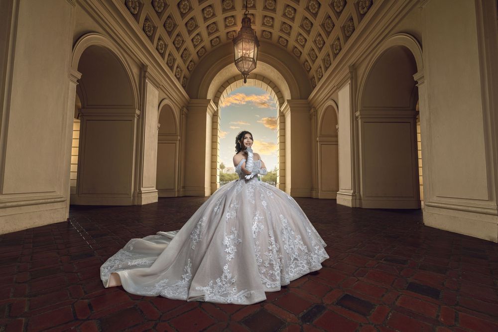Bride in elegant white ball gown with lace details stands in ornate archway hall with chandelier and colorful sky backdrop.
