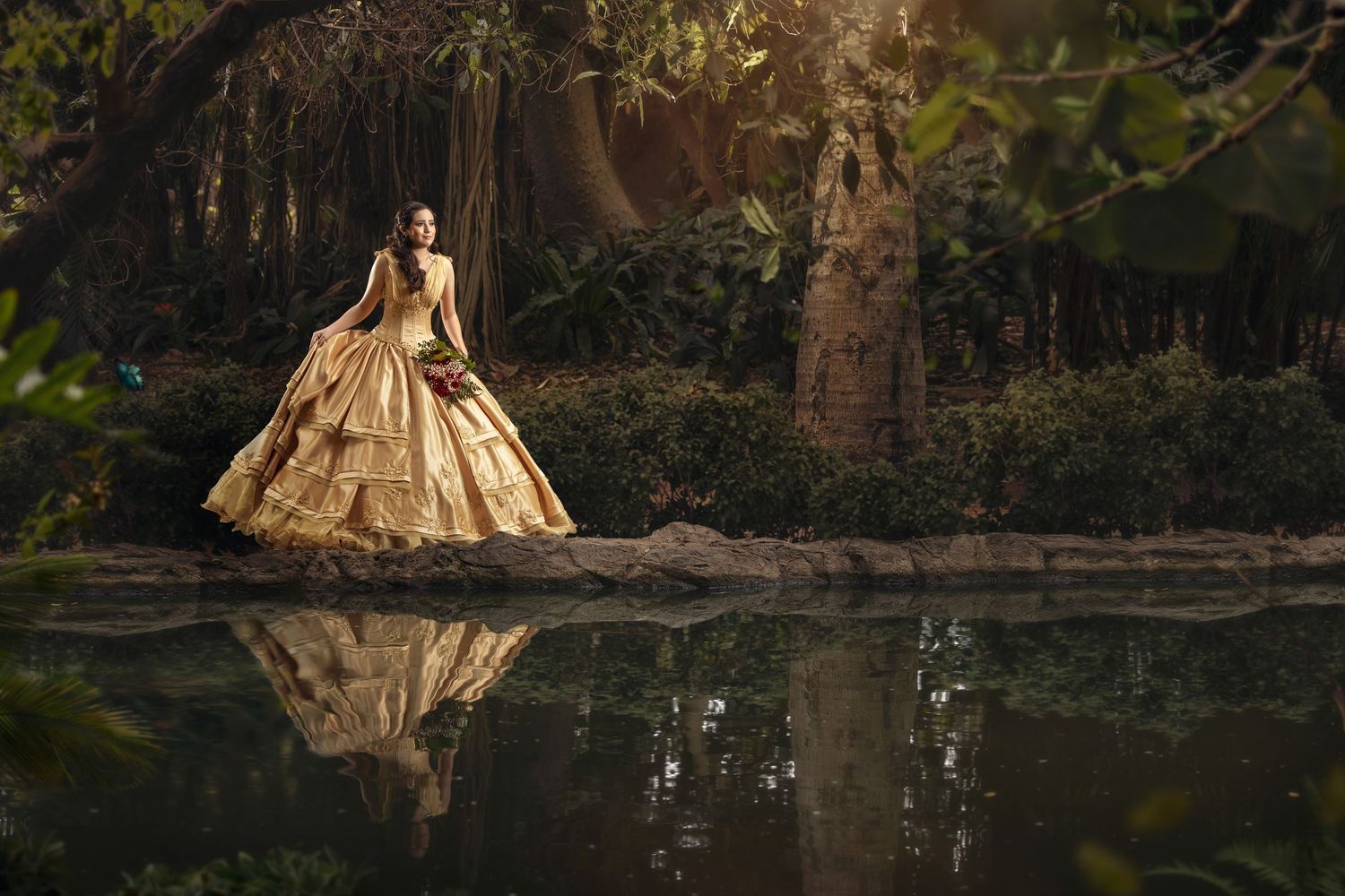 A golden vintage ball gown reflects in a tranquil pond surrounded by moody forest scenery at sunset.