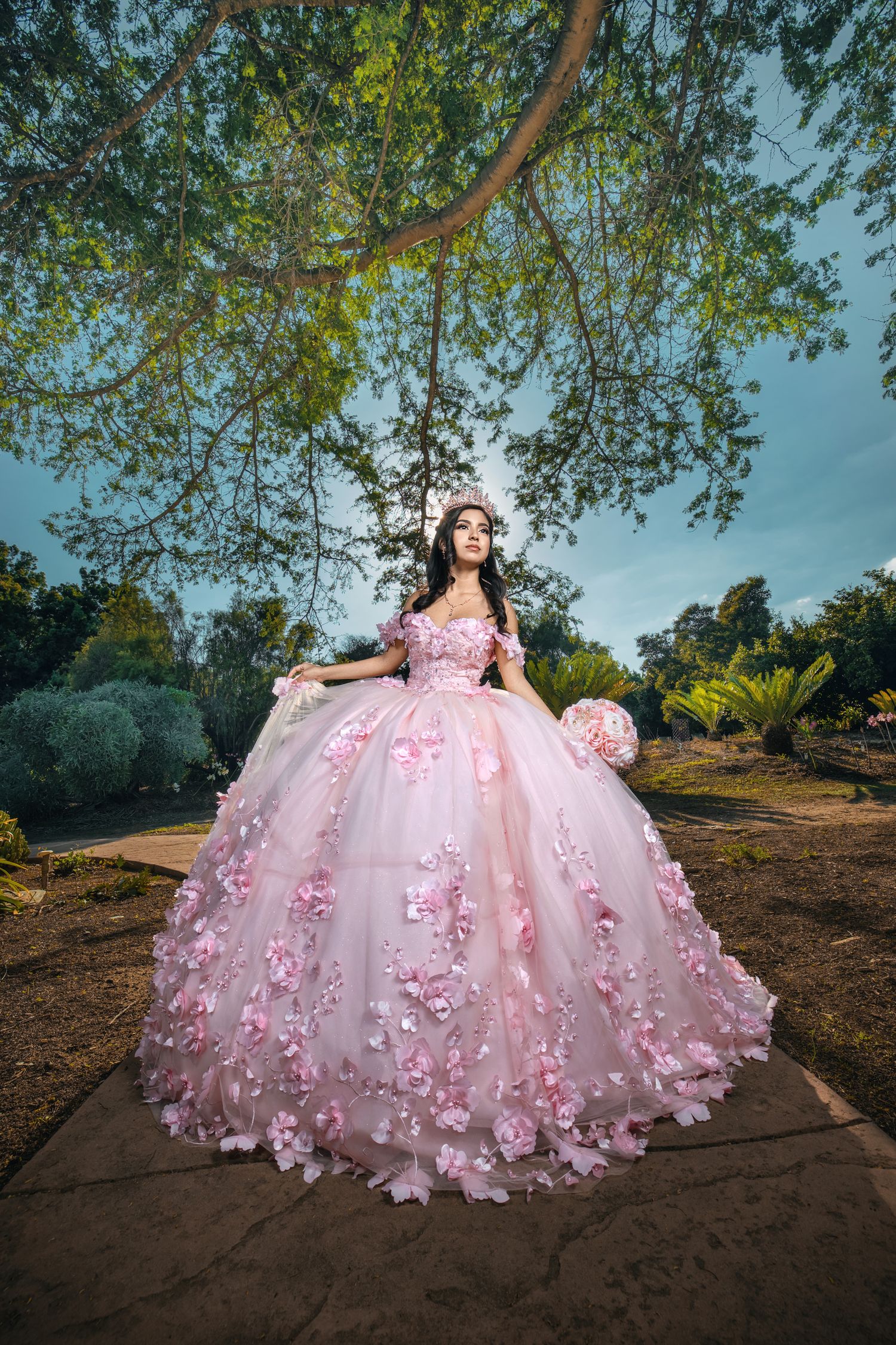 A stunning pink ball gown with floral appliques photographed in a garden setting under hanging tree branches.