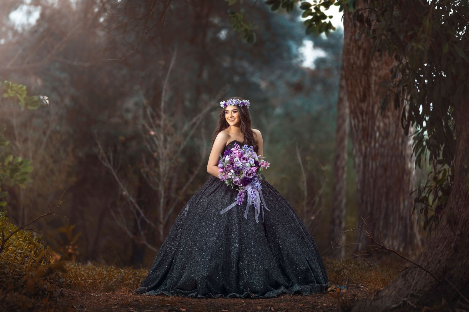 A person in an elegant black ball gown with purple flowers stands in a misty forest setting at twilight.