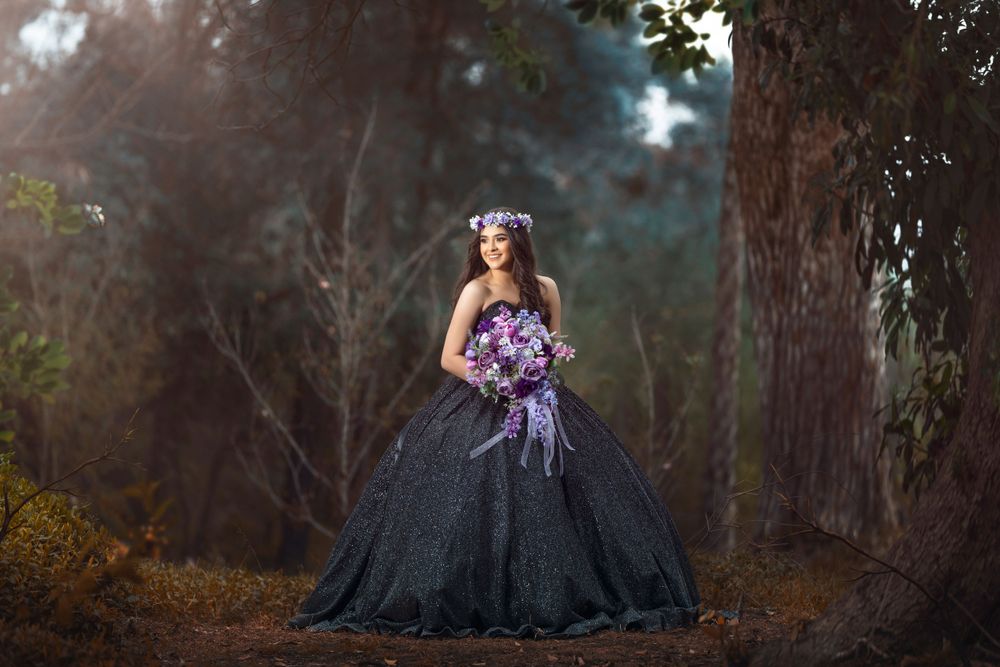 A person in an elegant black ball gown with purple flowers stands in a misty forest setting at twilight.