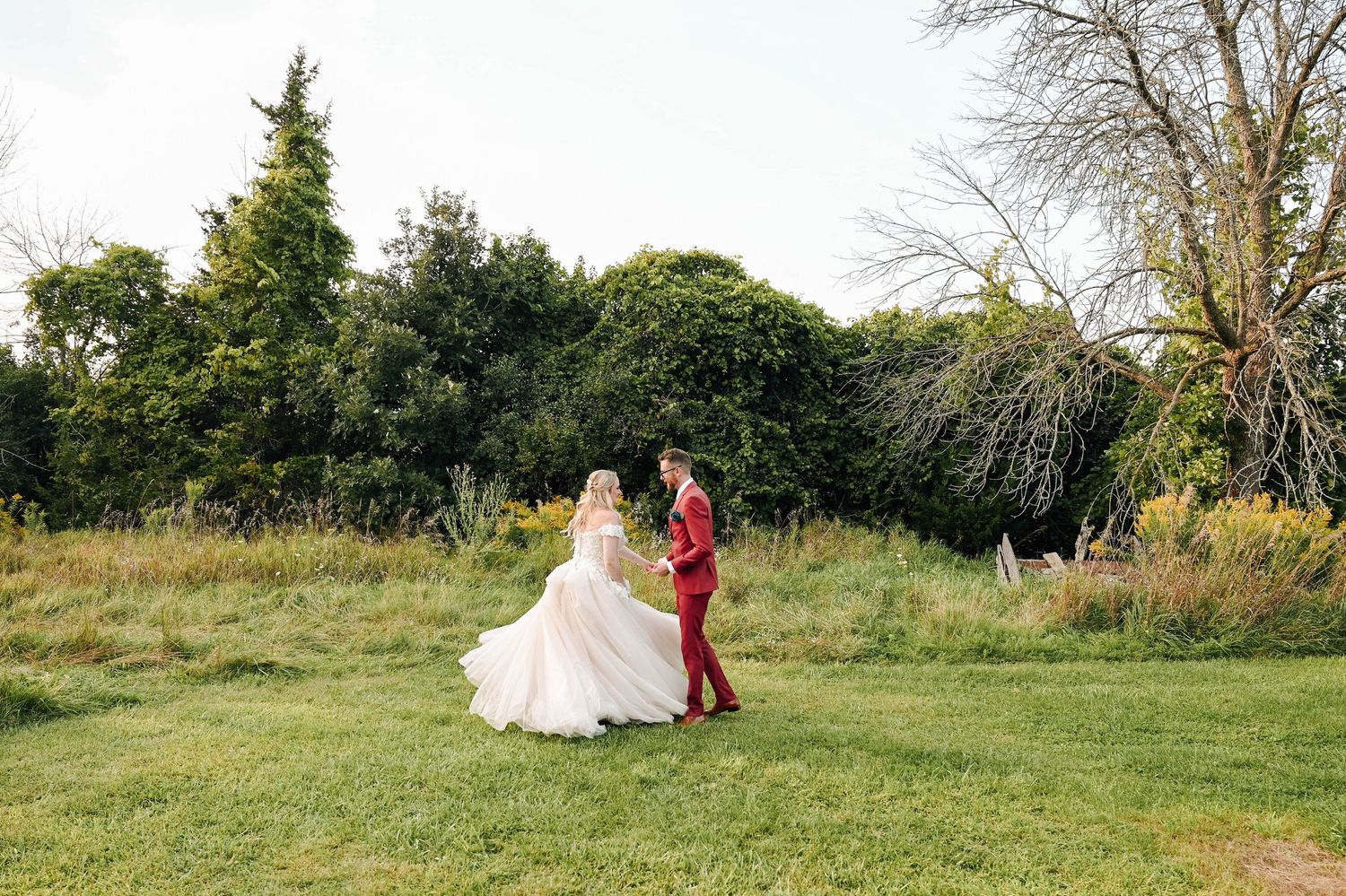 A couple in wedding attire dance together on a grassy lawn surrounded by lush summer greenery.
