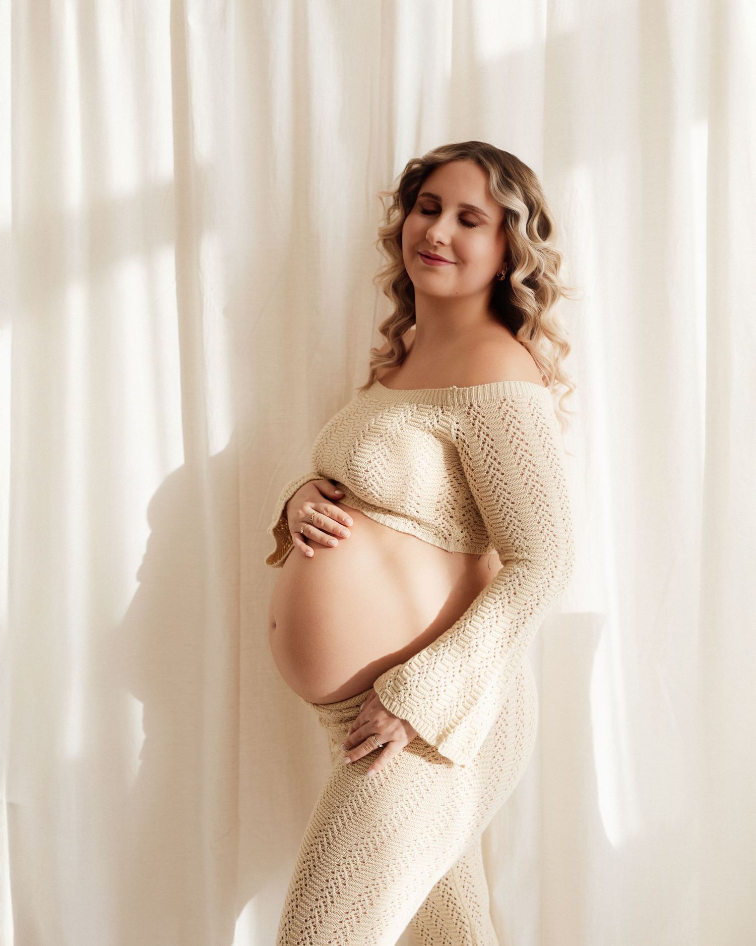 Pregnant woman draped in white fabric cradling her bare baby bump against a brown background.
