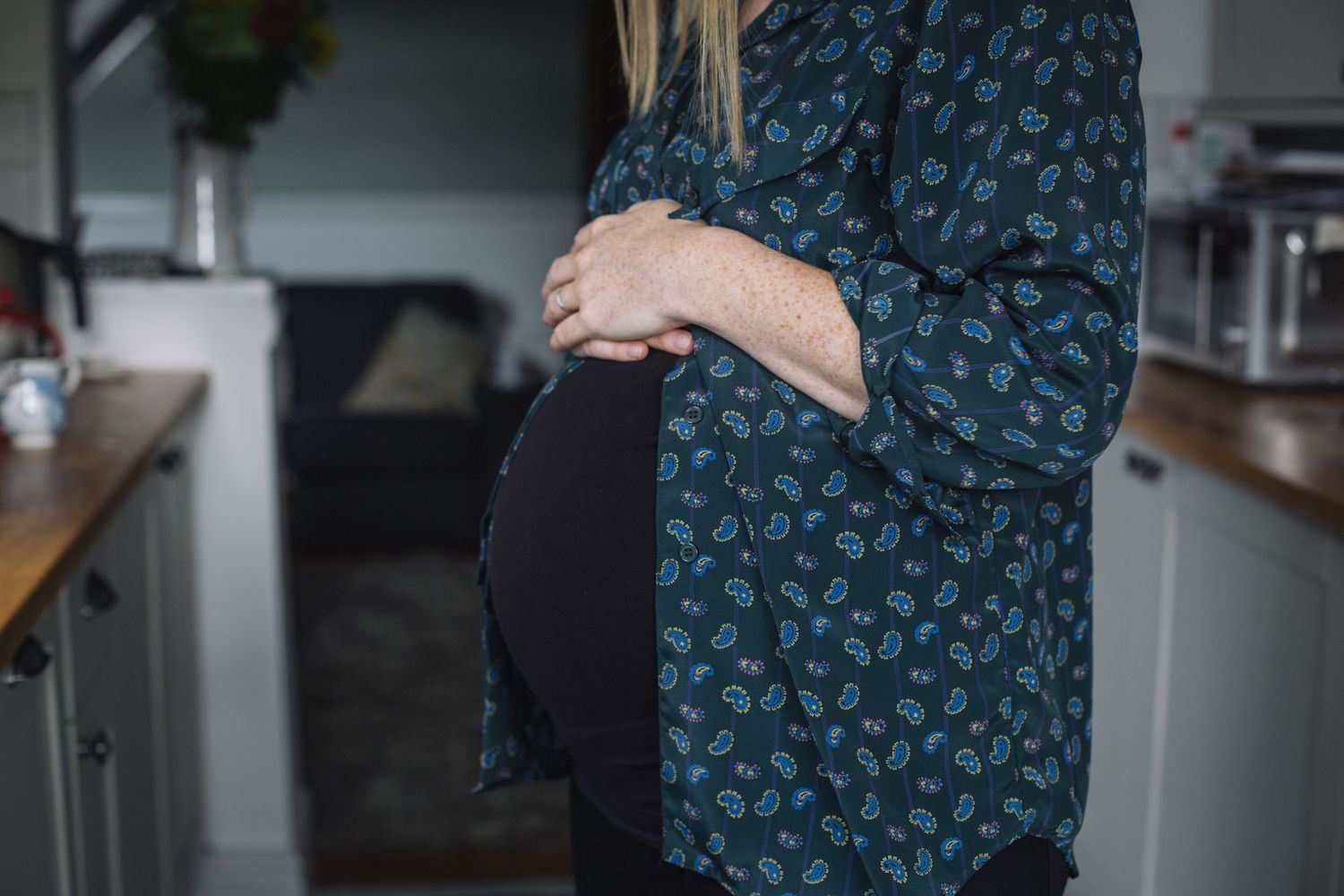 Side view of a pregnant belly being cradled by hands, wearing a dark patterned long sleeve shirt in a kitchen setting.