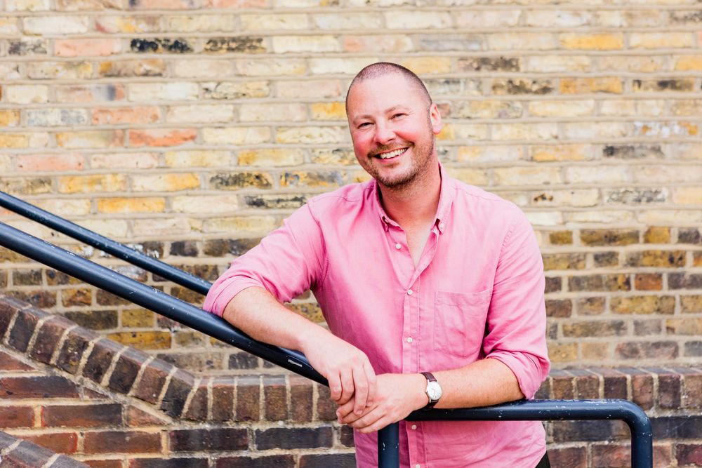 Portrait against brick wall wearing pink button-down shirt and casual attire.