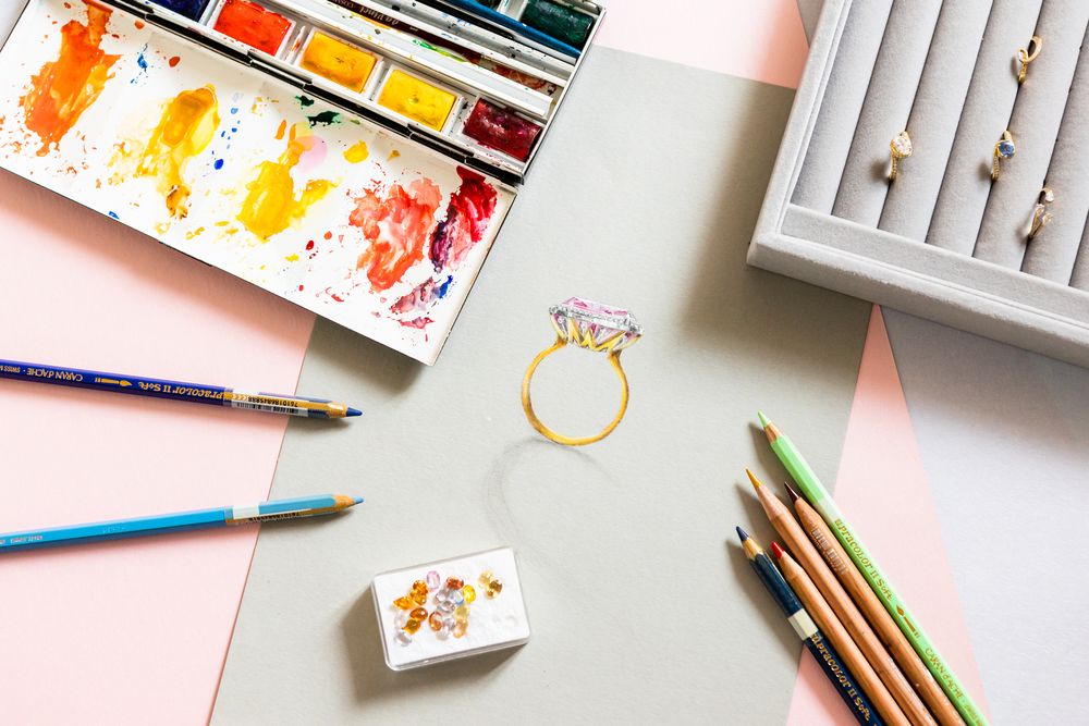Art supplies and watercolor palette with gold ring sketch on white paper.
