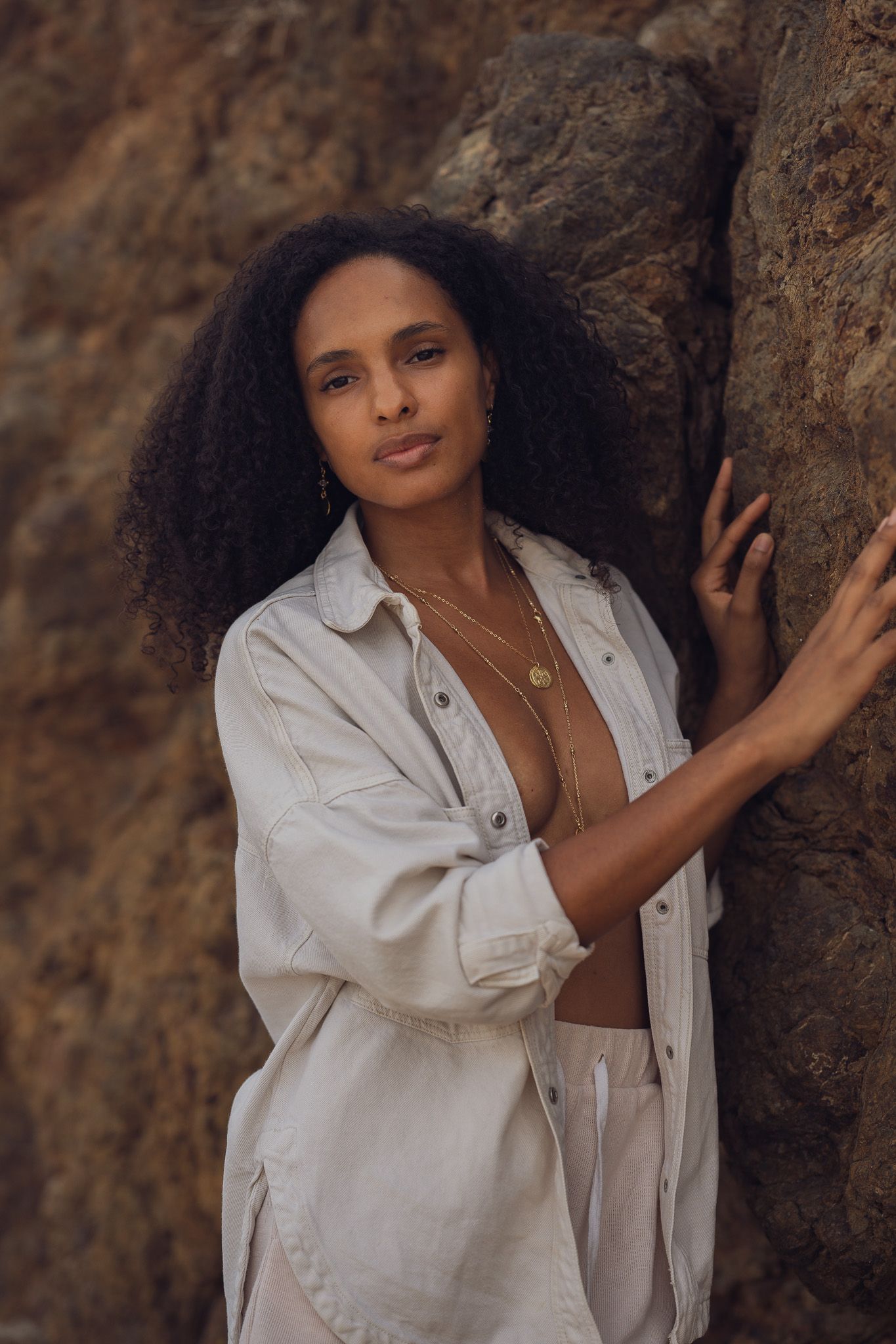 A stylish portrait of someone in a white button-down shirt posing against a textured rock wall in warm lighting.