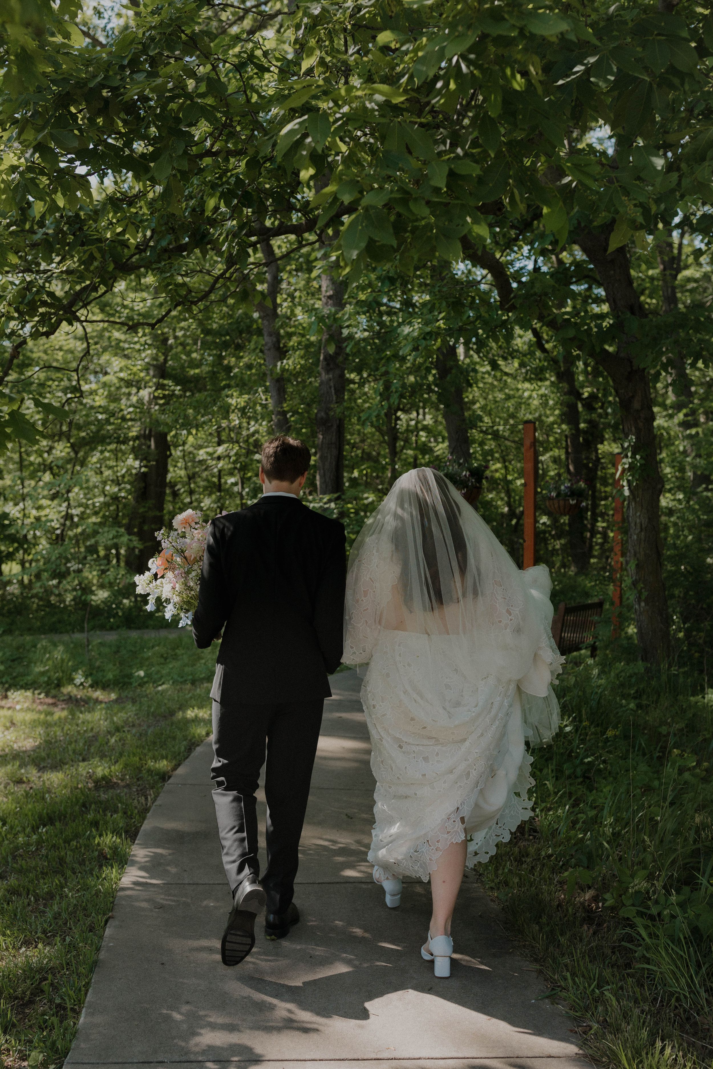 A bride and groom walk down a tree-lined pathway, with the bride's veil and dress trailing behind them.
