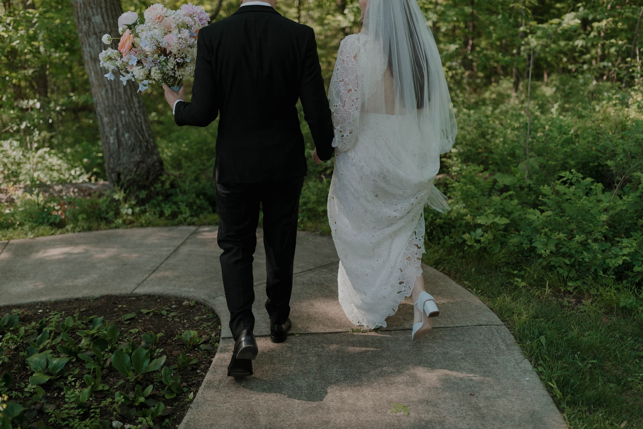 A couple in wedding attire walking together on a garden path during their outdoor ceremony.