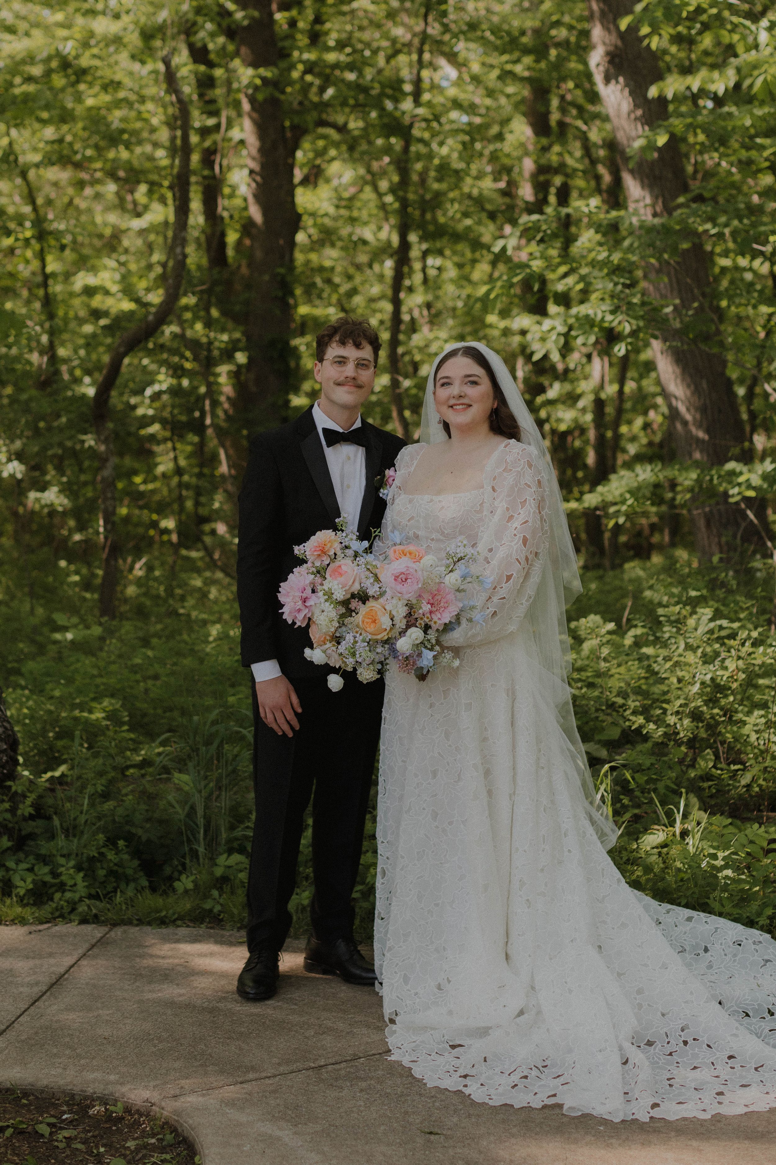 A wedding couple poses on a garden path surrounded by lush greenery and holding a pastel floral bouquet.