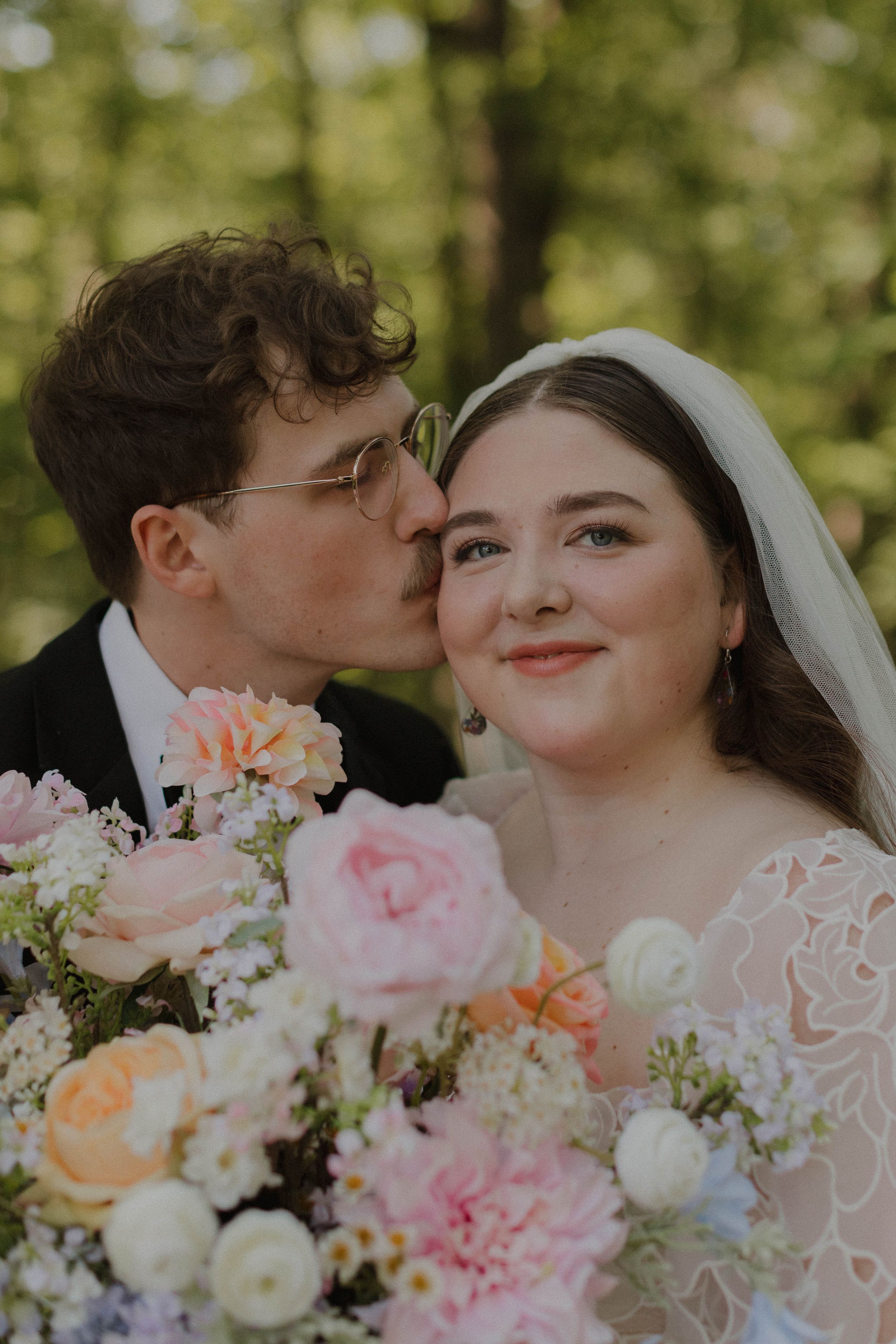A romantic wedding couple shares intimate moments while holding a bouquet of pink and white flowers in an outdoor setting.