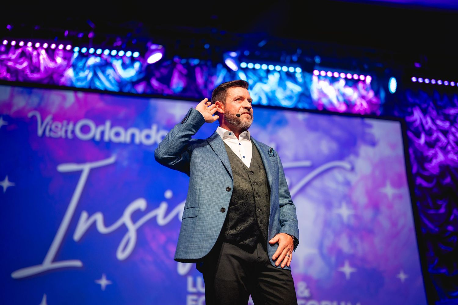 A performer in a light blue suit jacket and vest sings on stage with illuminated Visit Orlando signage in background.