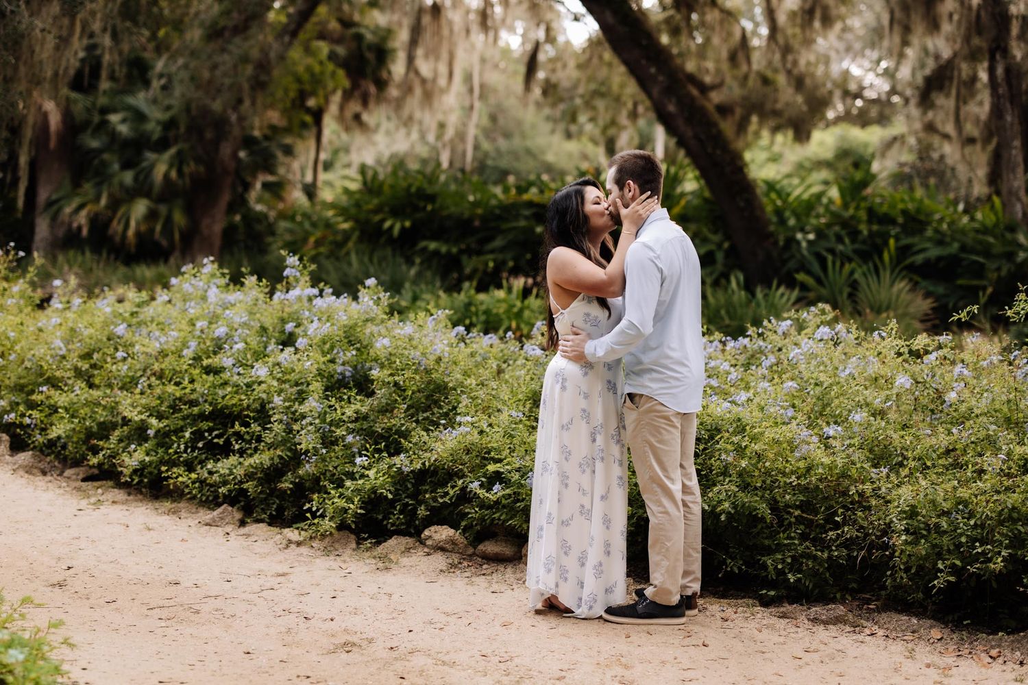 A romantic couple embraces near a wooden gazebo and pond in a lush garden setting at sunset.