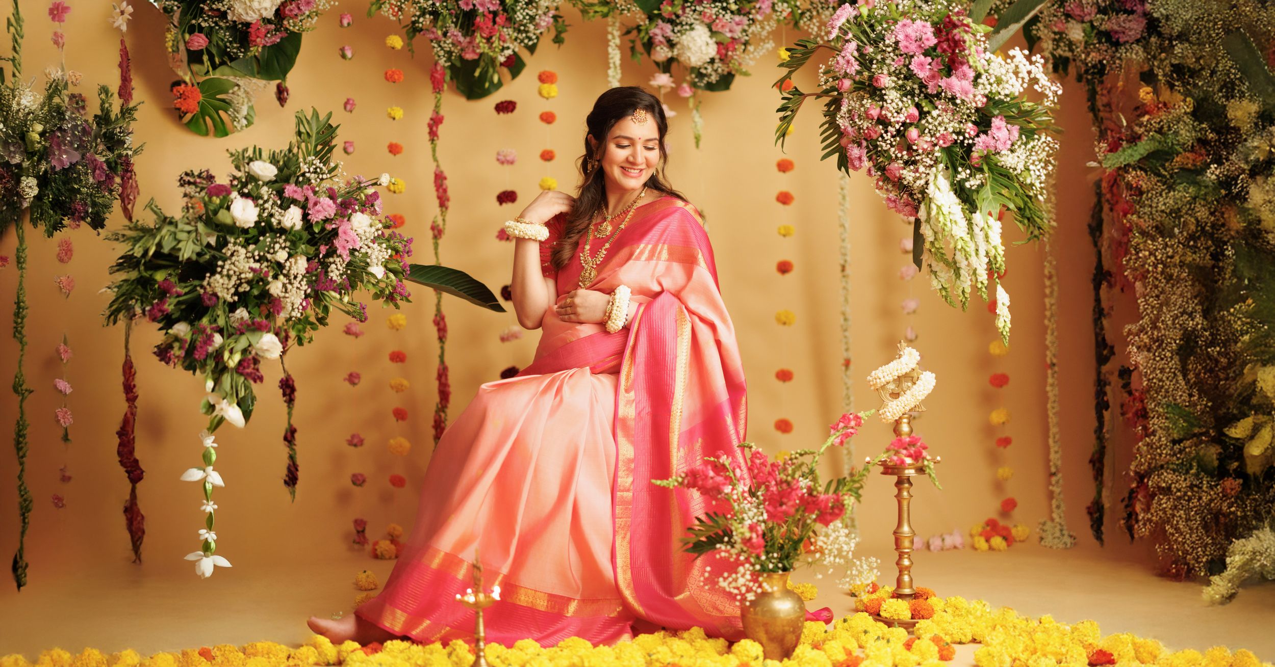 Traditional saree-clad figure poses elegantly against golden backdrop with hanging floral decorations and rose petals.