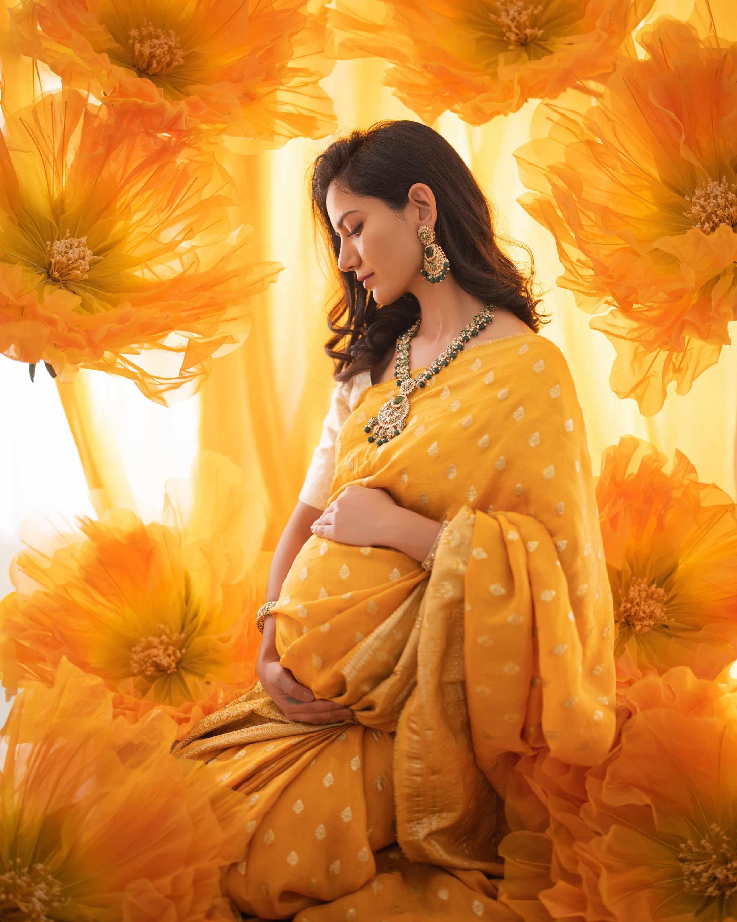 Maternity portrait against orange floral backdrop with figure in yellow sari adorned with jewelry in warm glowing light.