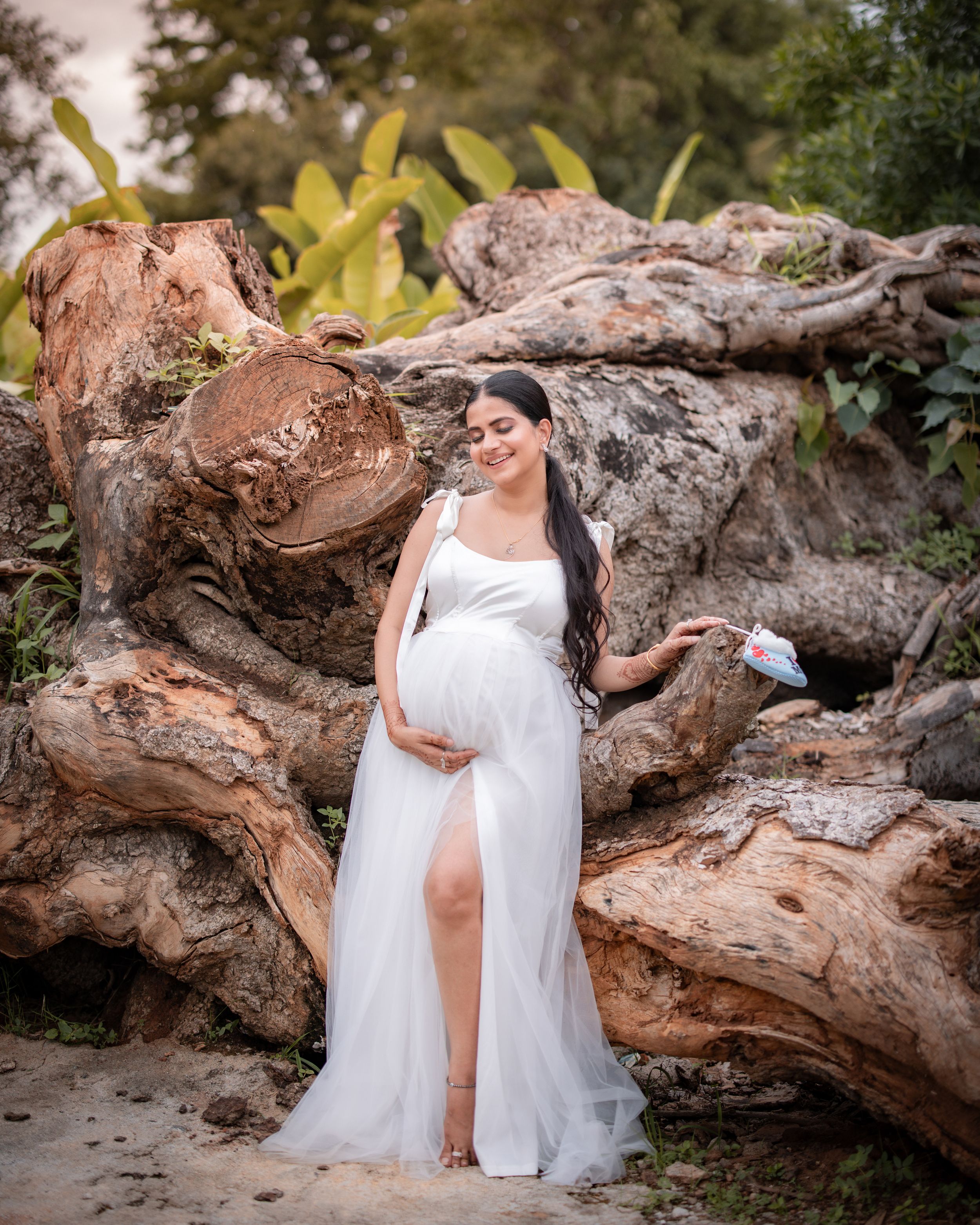 White flowing maternity dress against natural driftwood logs creates an ethereal beach portrait setting.