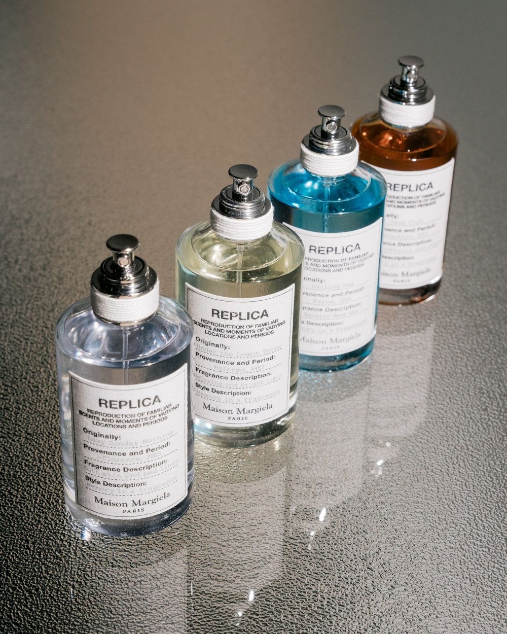 Four glass bottles of Replica fragrances arranged in a diagonal line on a reflective surface.