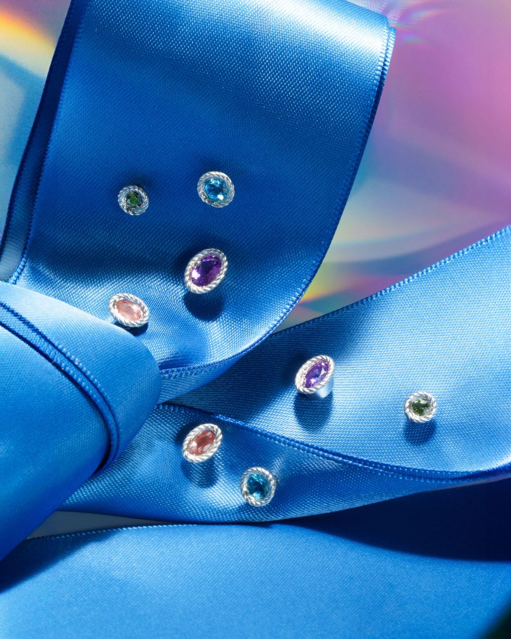 Colorful gemstones scattered across flowing blue satin ribbon creating an elegant jewelry display.