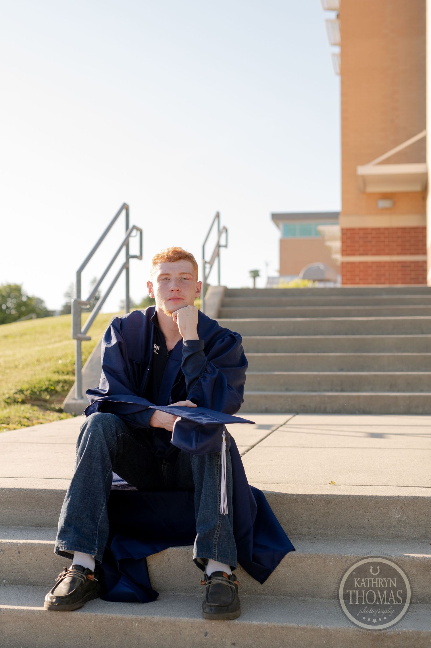 Graduate in navy gown sits contemplatively on concrete steps outside a brick building.