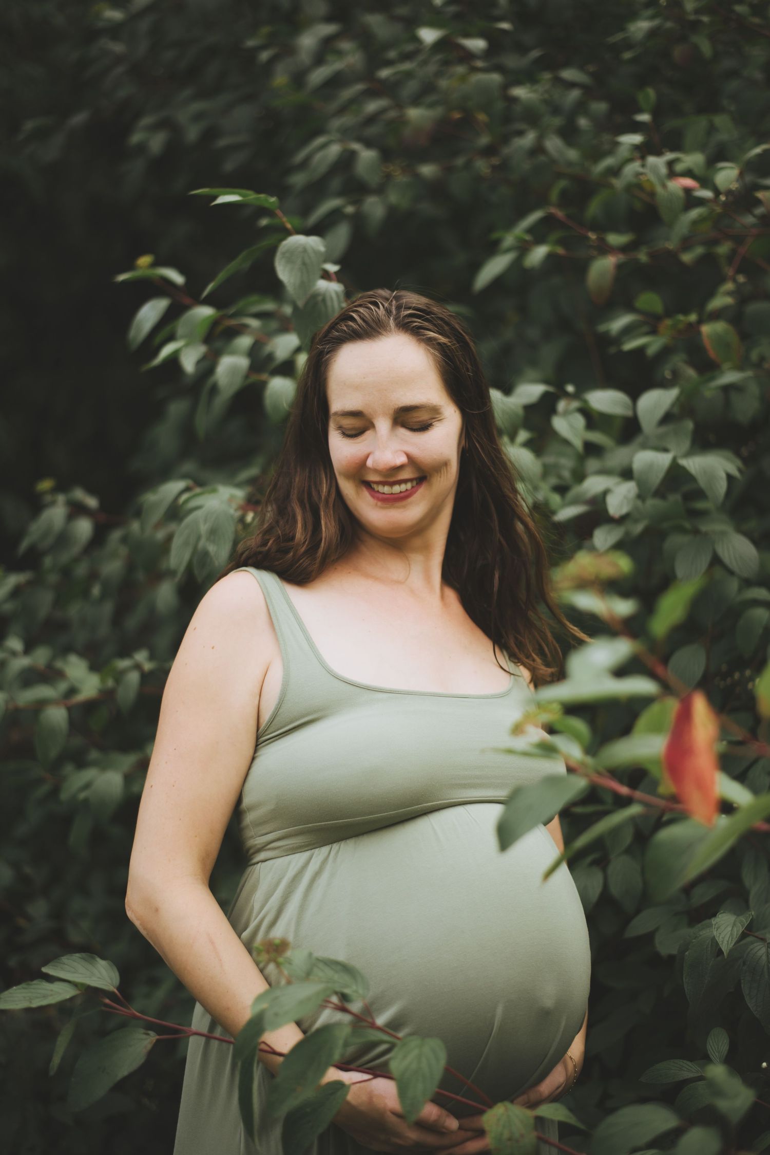 A pregnant woman in a sage green dress stands among leafy foliage in a natural outdoor setting.