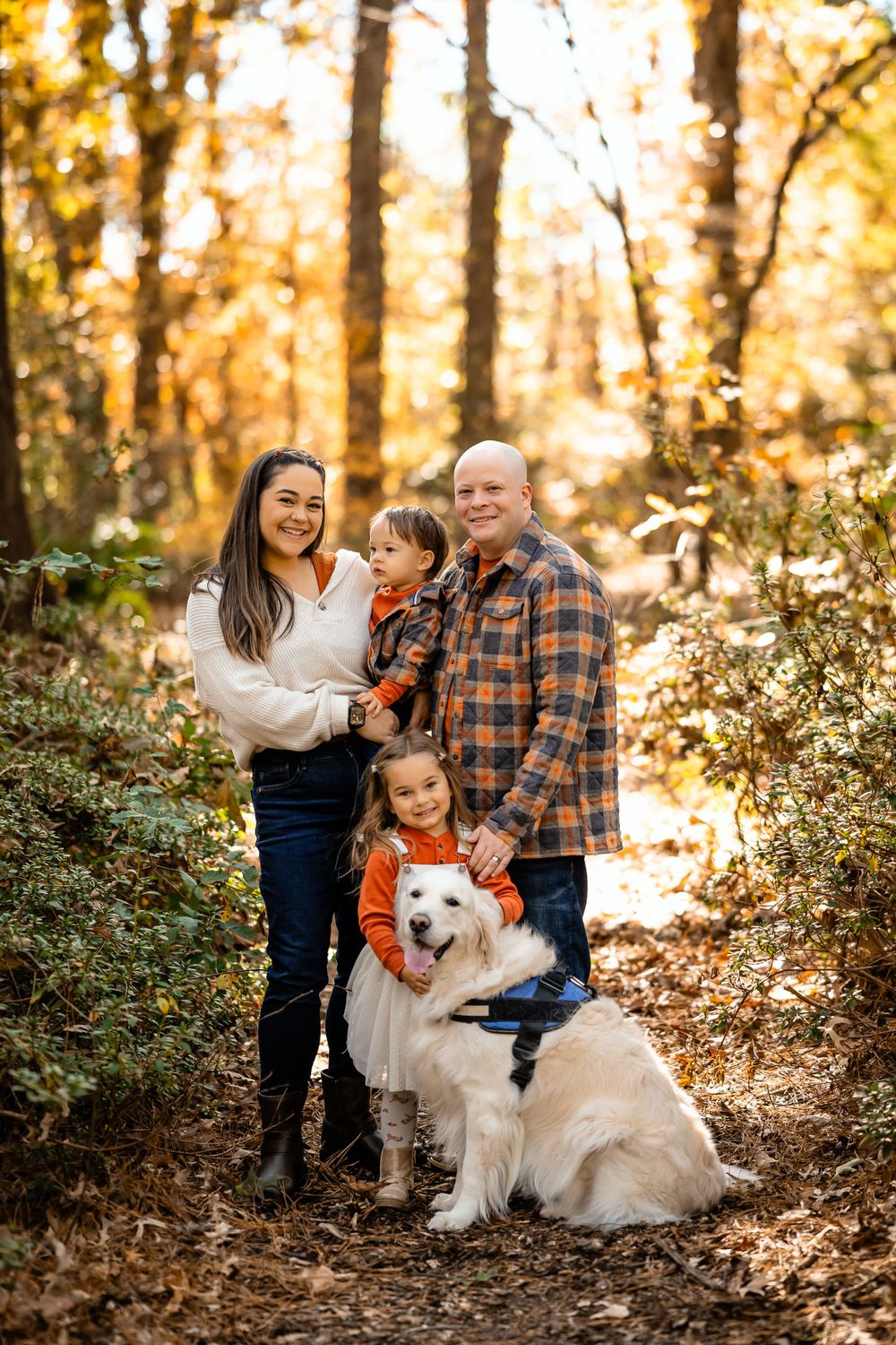 Family poses with their fluffy white dog on a wooded trail during fall season.