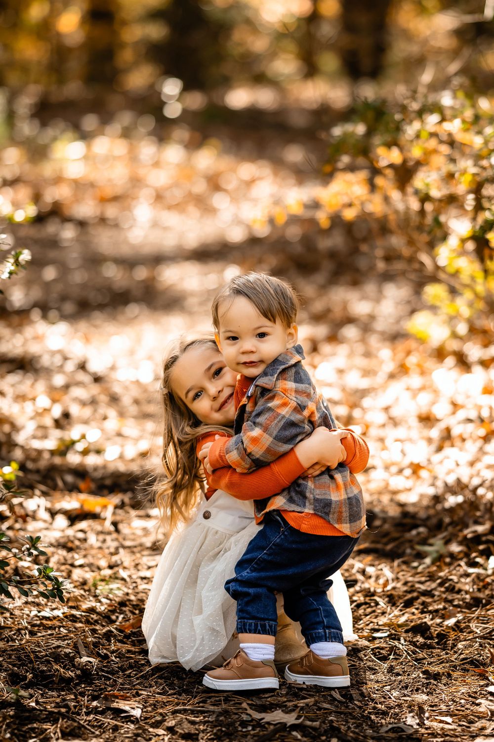 Young children hugging on leaf-covered forest path during autumn photoshoot.