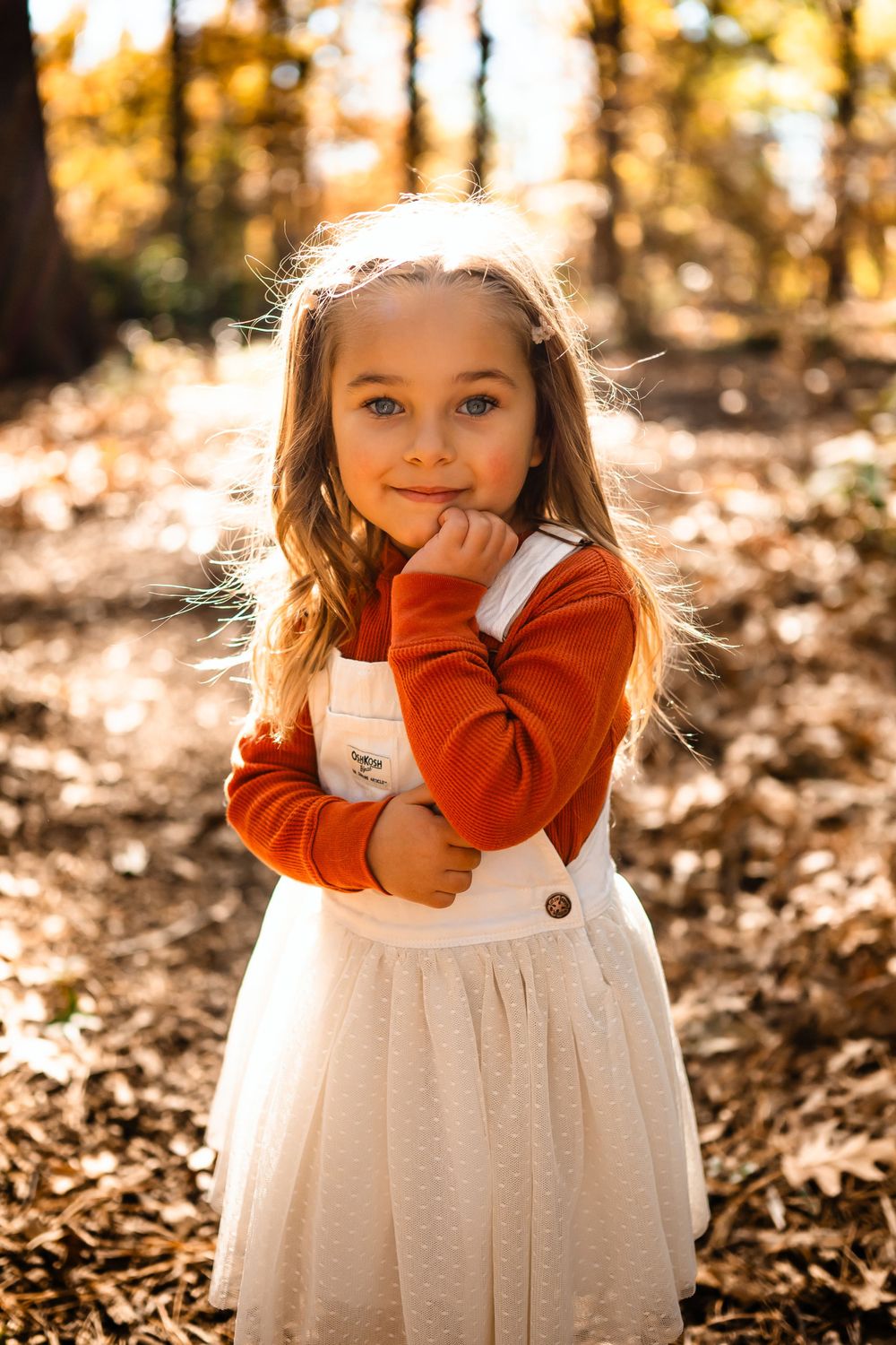 Young child in white dress and orange sweater poses in autumn forest setting.