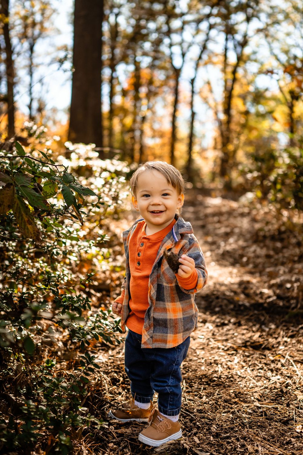 Toddler in orange and plaid shirt smiling while walking through autumn woods.