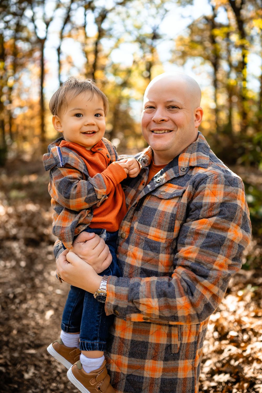 Adult holding smiling toddler while wearing matching plaid flannel shirts in forest setting.
