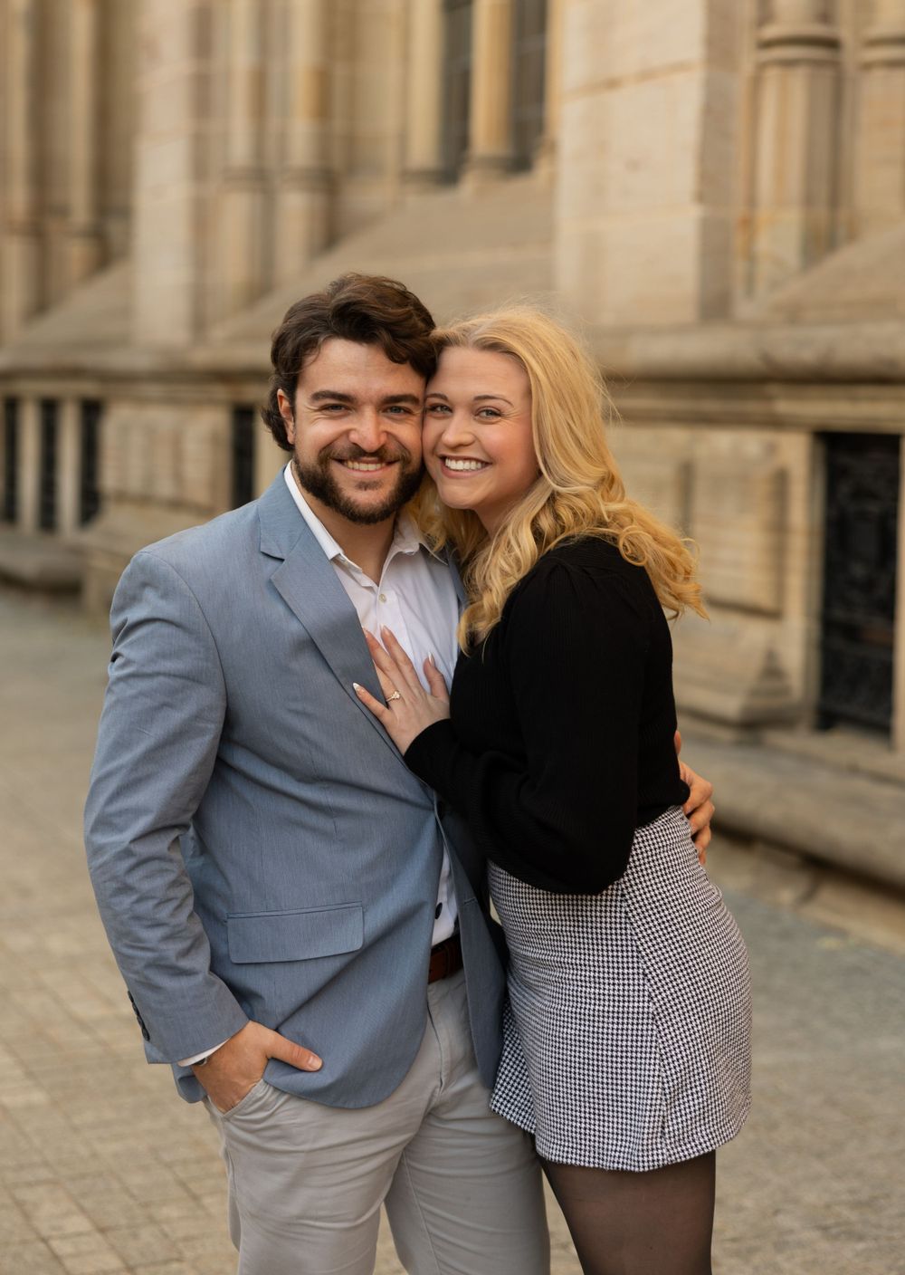 A romantic couple embraces and smiles together outside a classical stone building.