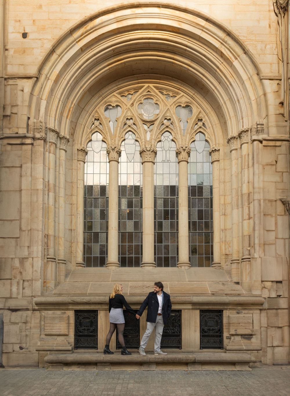 A couple poses against gothic church windows and stone architecture.