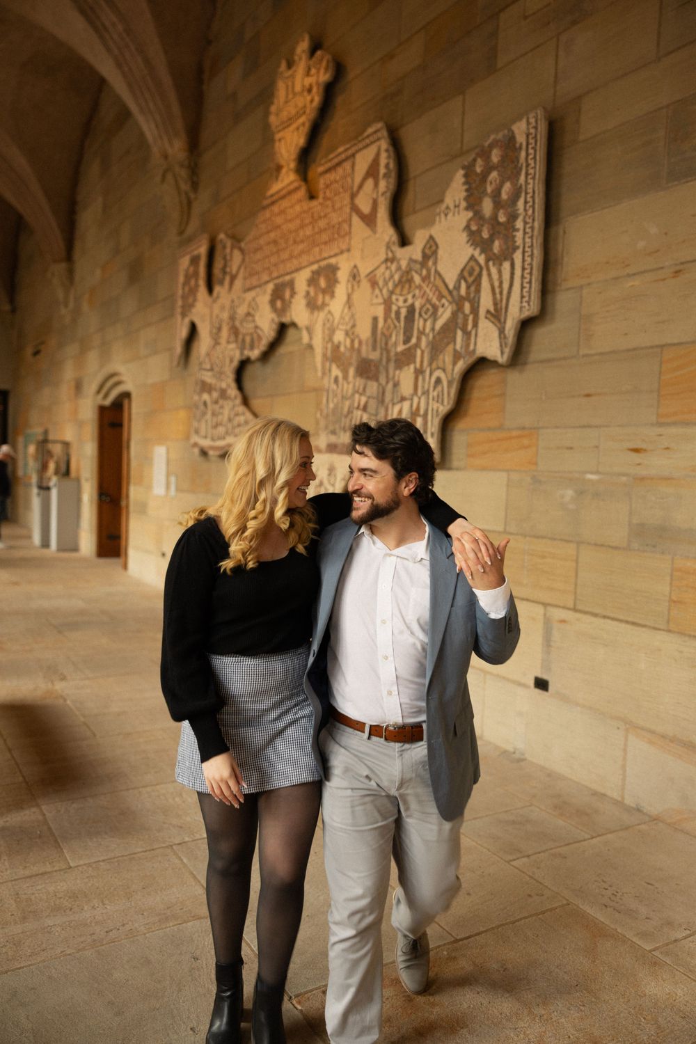 A couple walks together along an ornate stone hallway with decorative wall carvings.