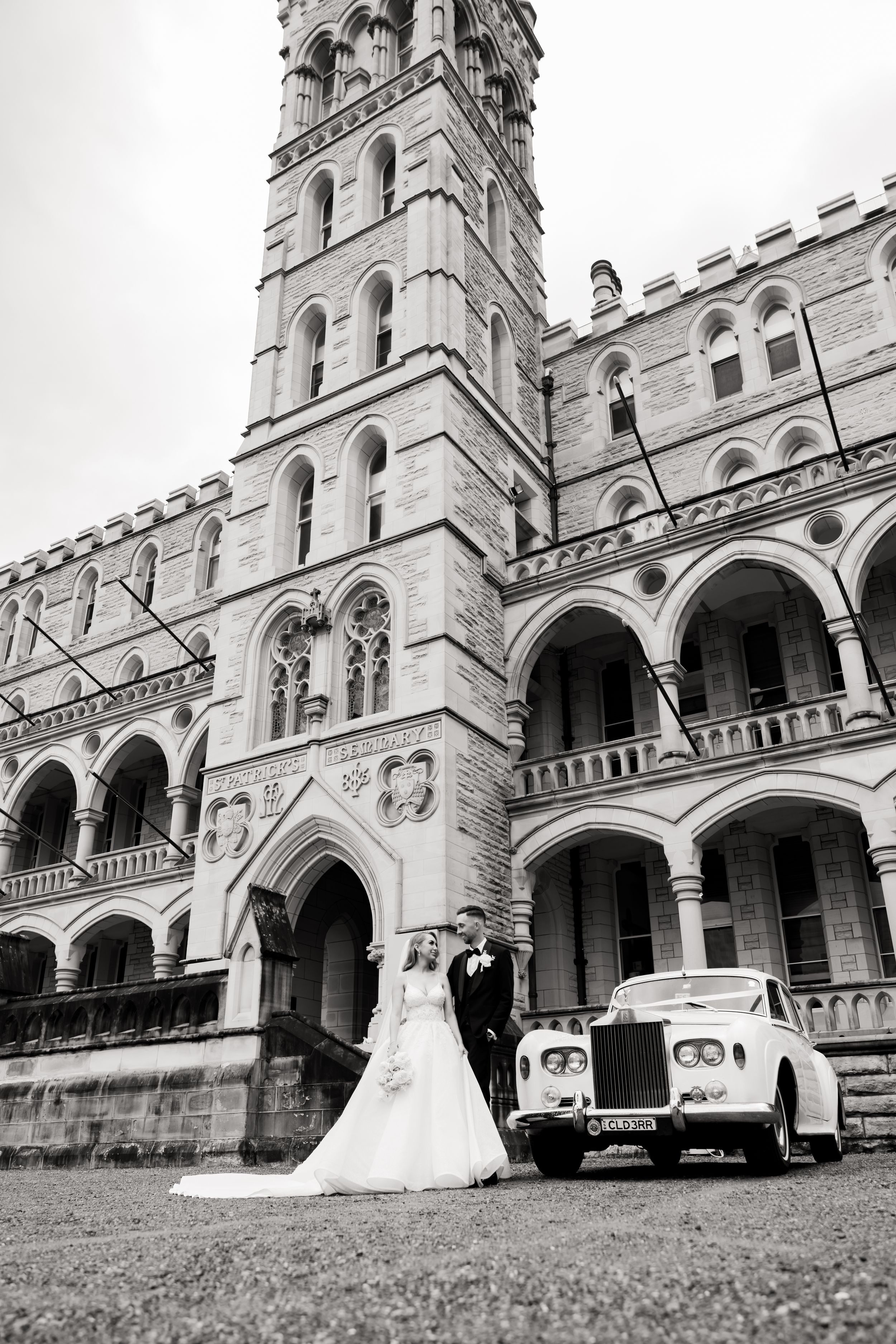 Wedding couple posing with vintage cars in front of a grand Gothic-style cathedral.