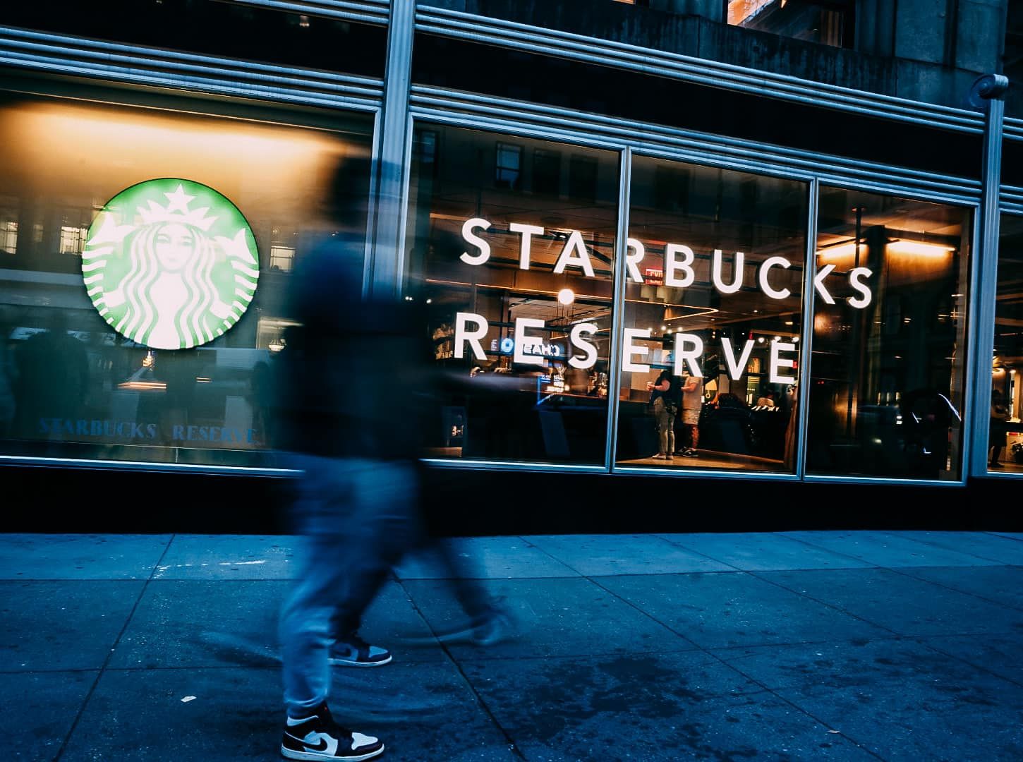 A blurred figure walks past an illuminated Starbucks Reserve storefront at night, with the iconic green mermaid logo visible.